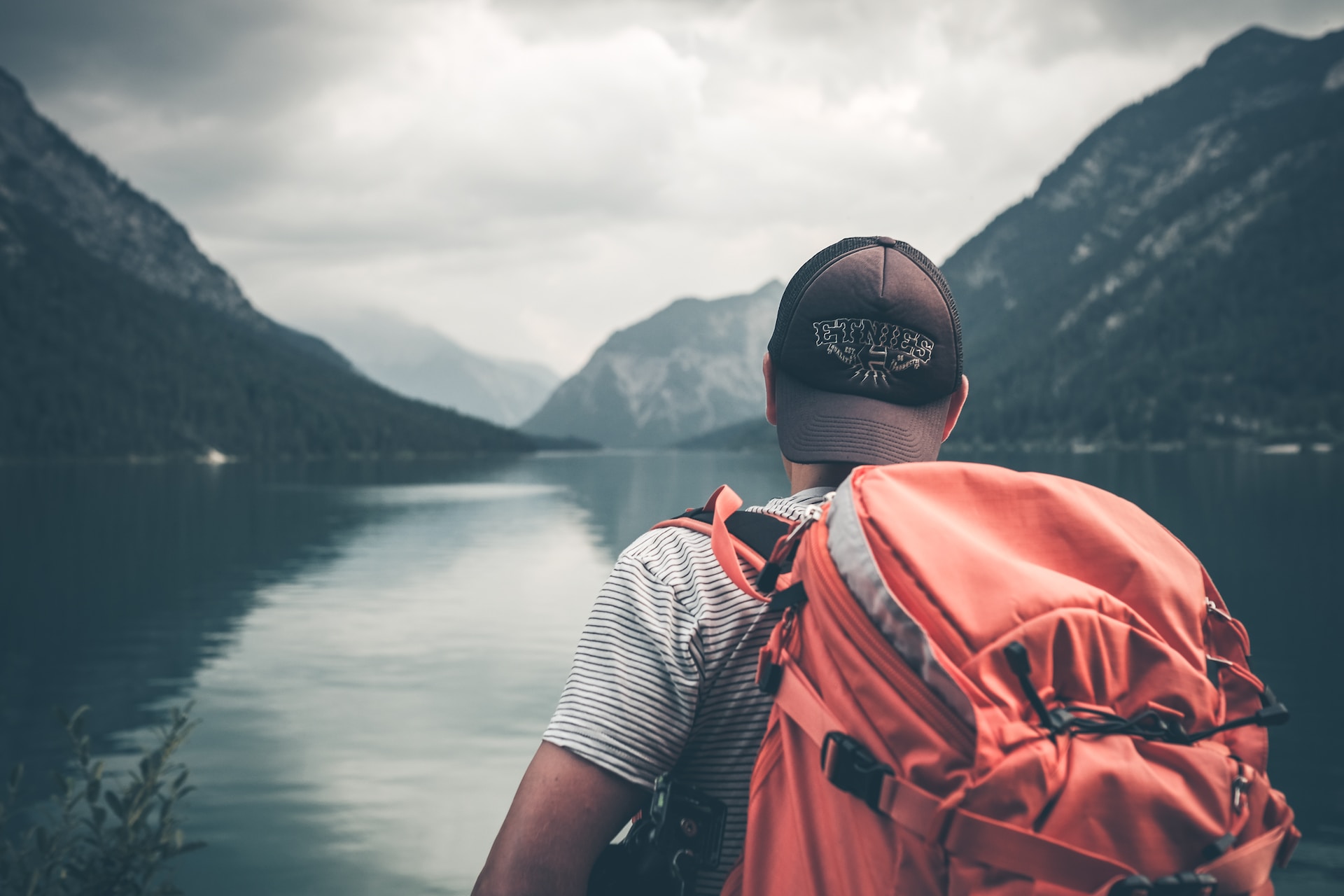 Man with a red backpack looking at a lake surrounded by tall mountains with a cap on backwards