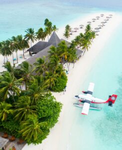float plane parked on a white sandy beach with palm trees and villas and clear water