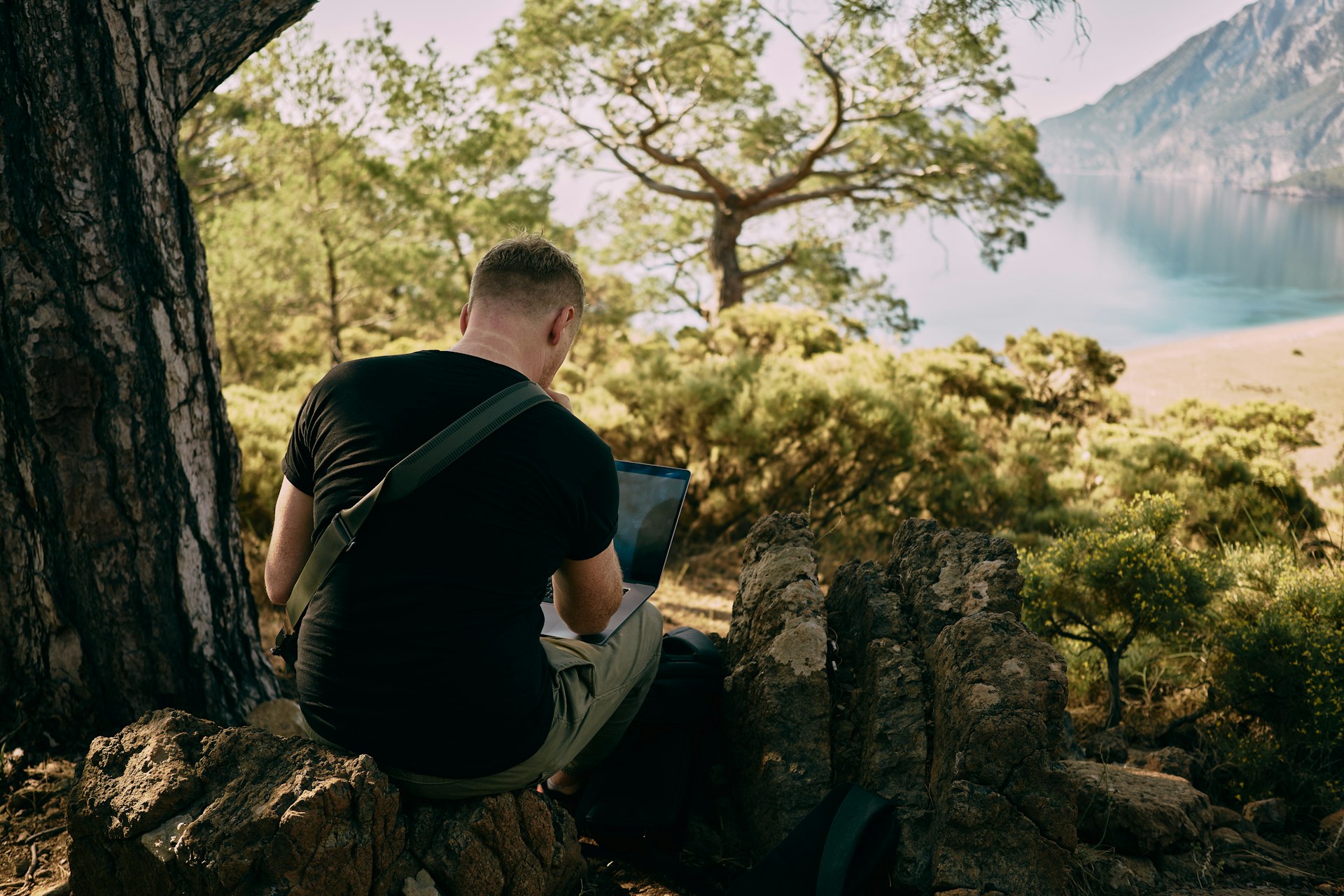 Guy working under a tree overlooking a lake with his laptop