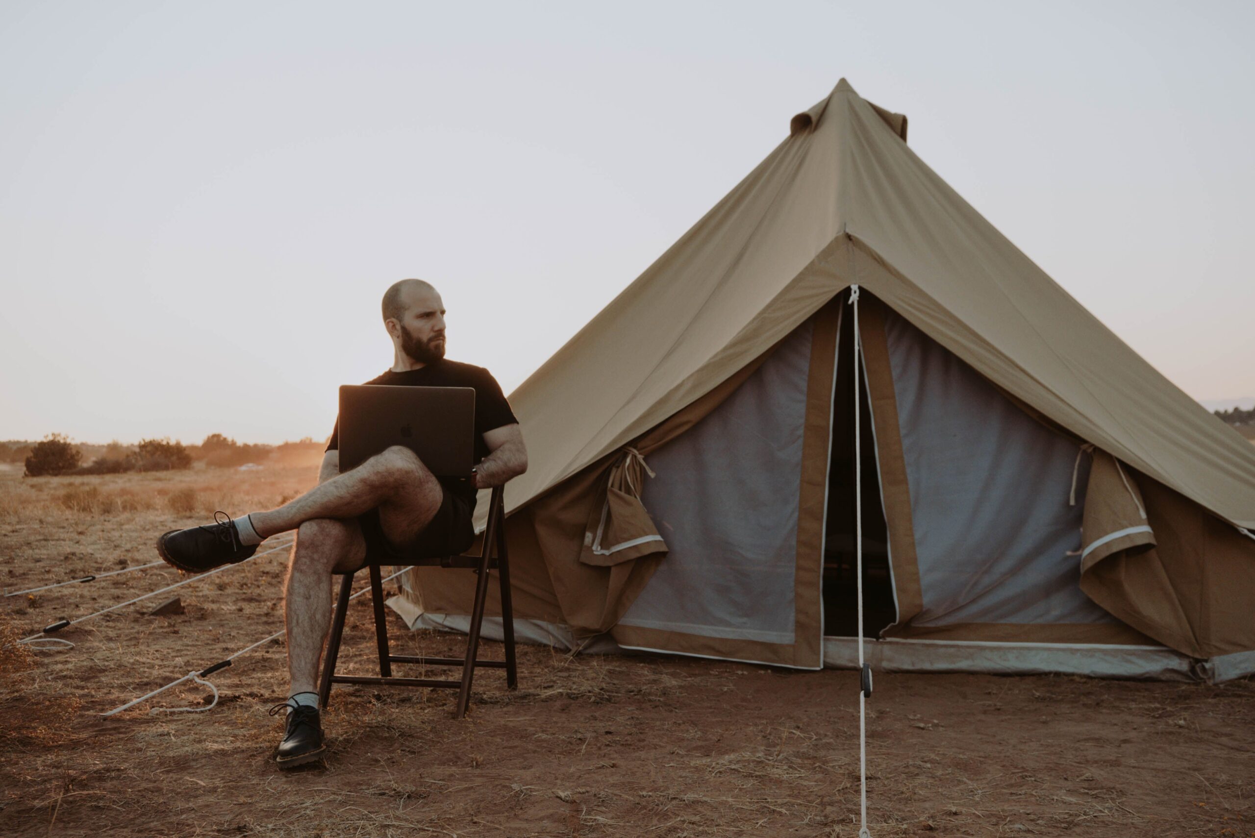 A digital nomad working on a laptop outside a bell tent at sunset, booking a stay while on the road
